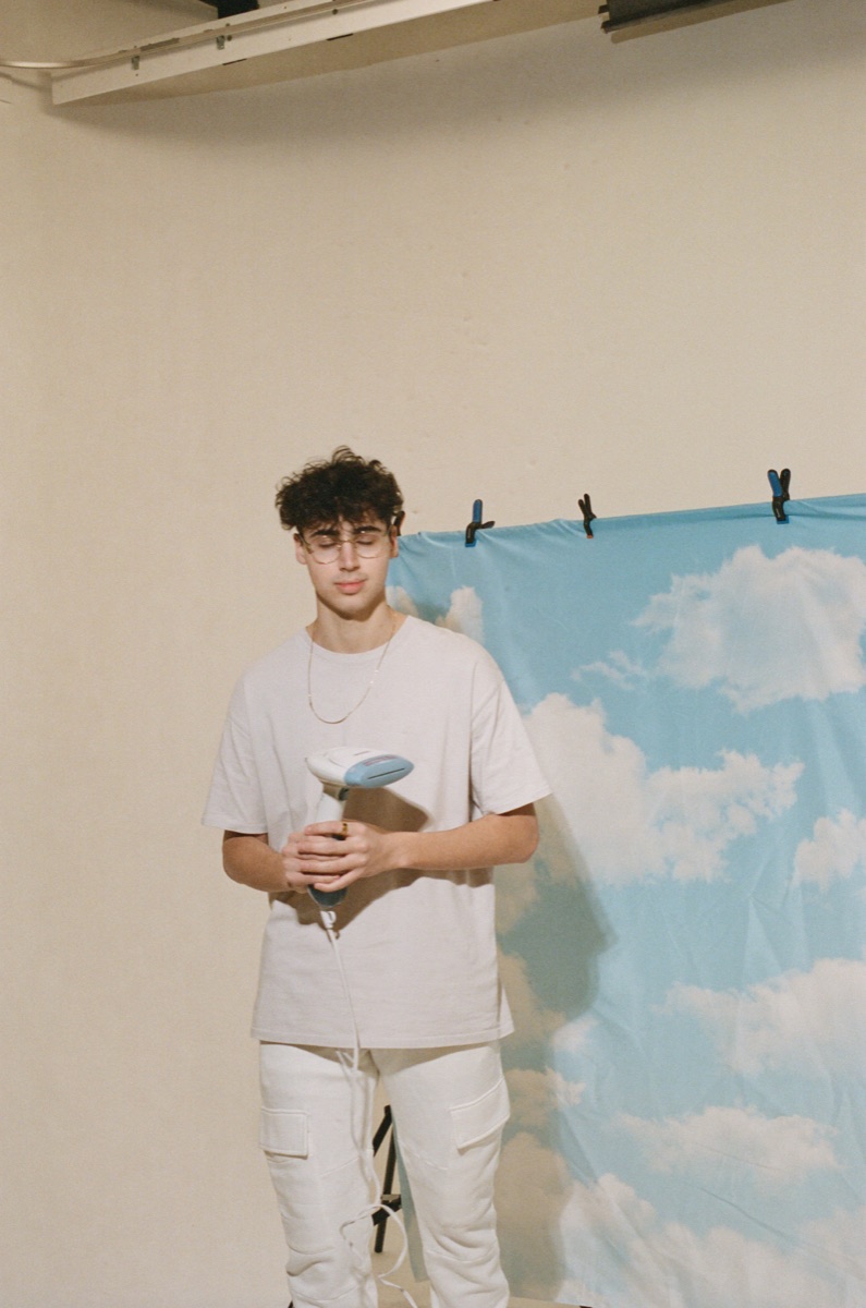 mgx standing under sky - Blog A young man stands quietly under a sky-and-clouds backdrop holding a cup, calm and reflective, in warm film grain