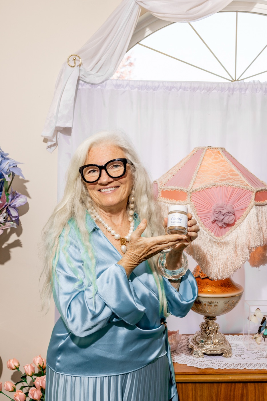 fifty plus woman window - Blog A woman with long grey hair and glasses standing by a bright window at home, holding a jar of capsules