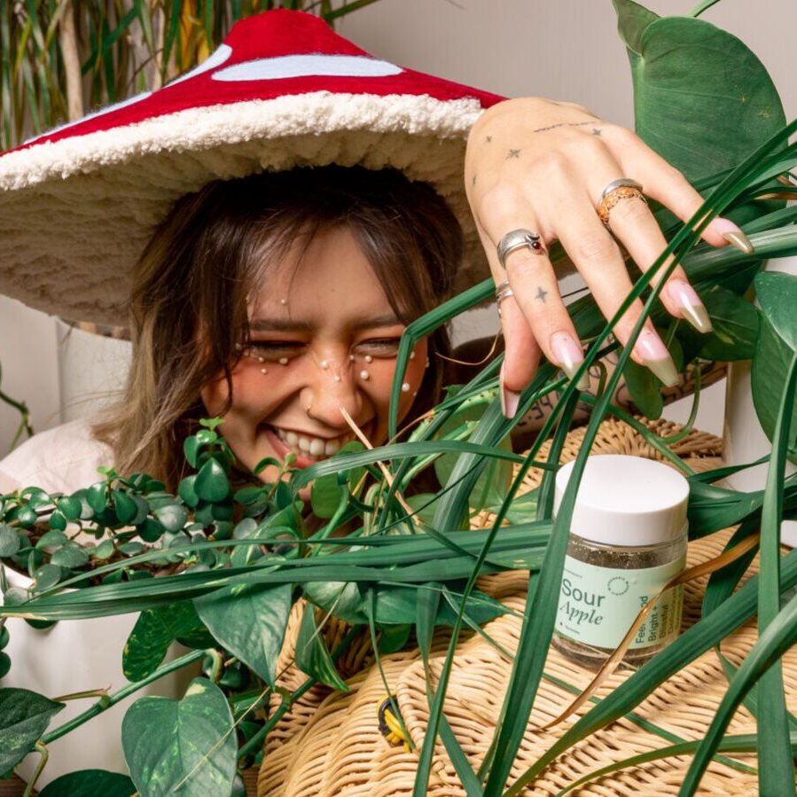A woman in a red mushroom hat laughing behind the plants, a jar of Sour Apple gummies beside her