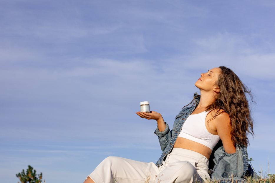 wp26 walk - Blog A woman sitting on a hilltop with her face turned up to the sun, holding a jar of Support Capsules